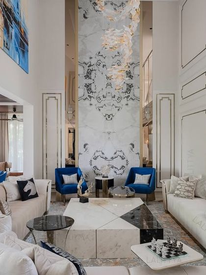 A wide view of the double-height living room, showing how we use symmetry to create a sense of order and grandeur. The two cobalt blue chairs provide a focal point against the neutral palette, anchored by a massive book-matched marble feature wall that draws the eye upward.