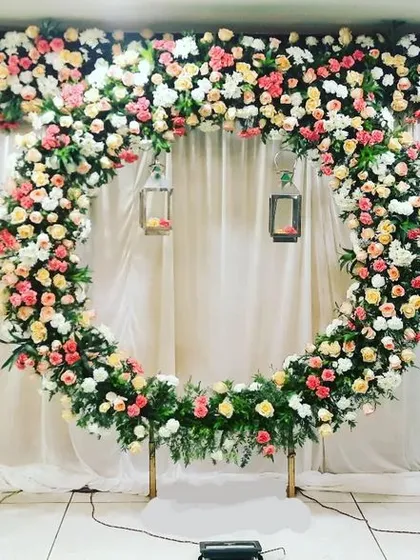 This engagement stage features a large, circular floral arch made of fresh roses, creating a stunning focal point. The setup is balanced with symmetrical floral arrangements on pedestals and a clean white curtain backdrop, perfect for an elegant indoor celebration.