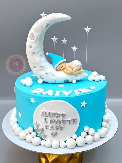 A "Happy 1 Month" celebration cake, with a tiny baby sleeping on a crescent moon.