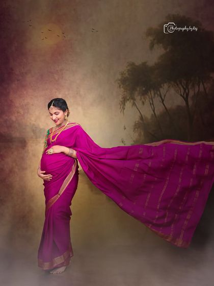A beautiful studio portrait of a mother-to-be in a magenta silk saree, with the fabric flowing gracefully. The painted backdrop adds an artistic touch.
