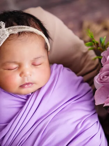 A close-up of a sleeping newborn wrapped in a lovely lavender swaddle, adorned with a floral headband and surrounded by matching flowers.