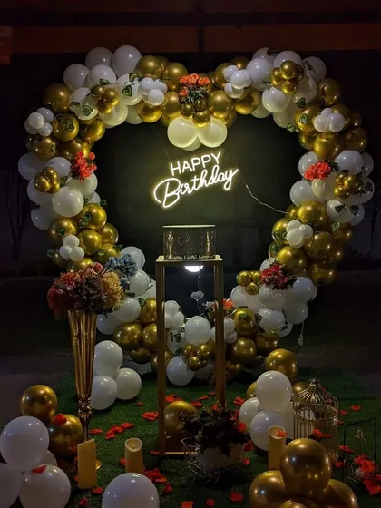 A glamorous birthday backdrop featuring a large heart-shaped balloon arch in gold and white. A "Happy Birthday" neon sign glows in the center, with a pedestal for the cake, all set for a memorable celebration.