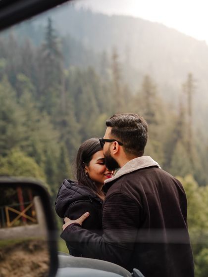 A creative shot capturing a tender forehead kiss, framed by the car's side mirror, with the Manali forest in the background.