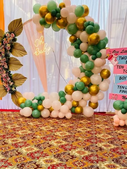 This gender-neutral baby shower decor features a half-ring frame decorated with green, white, and gold balloons. An "Oh Baby" neon sign and fun gender-reveal signs on the side add a playful yet stylish element to the party.