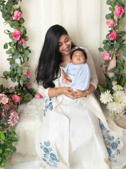 A mother smiles at her peacefully sleeping newborn, wrapped snugly in a blue swaddle, during their floral-themed studio session.