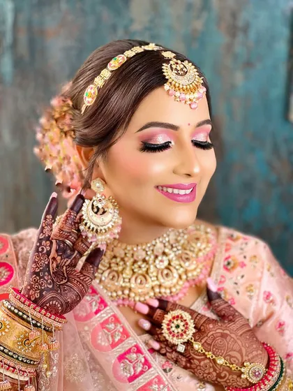 A joyful bride in pink, showcasing a beautiful Kundan and Polki set with a matching sheeshpatti. The intricate mehndi on her hands is beautifully displayed.