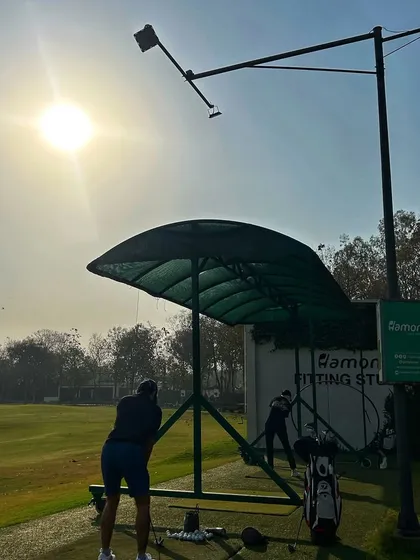 A golfer practices from a shaded bay, with the sun high in the sky and our Fitting Studio in the background.