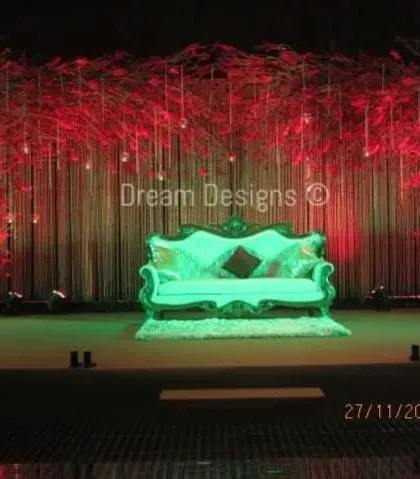 A stage design that plays with colored lighting for a dramatic effect. The backdrop of bare branches is washed in red light, while the central sofa is highlighted in a contrasting green, creating a bold and artistic look.