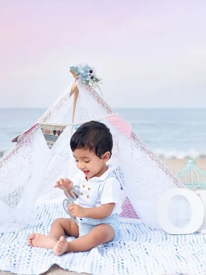 A baby boy celebrating his first birthday with a sailor-themed setup on the beach, complete with a teepee and "ONE" letters.