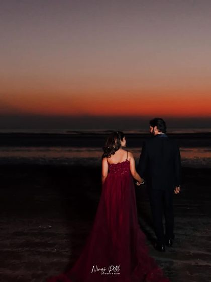A couple holding hands and walking on the beach at sunset. The bride-to-be wears a beautiful red gown with a long trail, creating a romantic and cinematic scene.