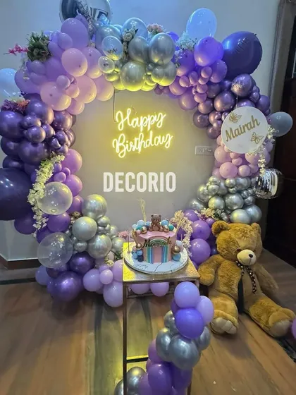 A teddy bear-themed birthday party with a gorgeous purple and silver color scheme. A large teddy bear sits beside the cake table, which is framed by a circular balloon arch and a neon sign.