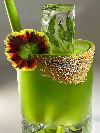 A vibrant green cocktail with a Tajin-dusted rim, garnished with a large, decorative leaf and a custom ice cube containing herbs. This drink is a masterpiece of color, texture, and flavor.