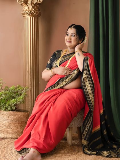 A beautiful solo portrait of a mom-to-be in a red and black saree, seated gracefully next to a golden pillar, looking radiant.
