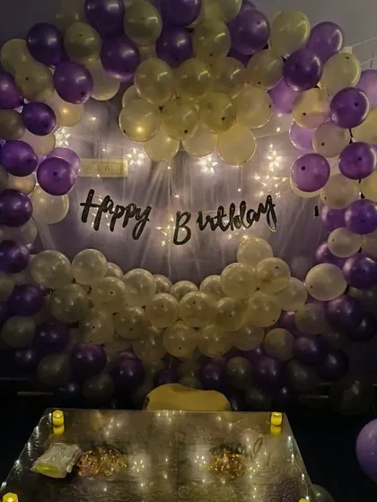 A simple yet classic birthday decoration. This circular balloon arch in purple and white is accented with fairy lights and a 'Happy Birthday' banner for a clean and festive look.