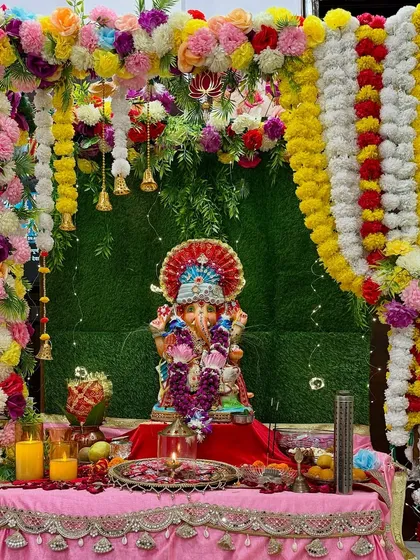 We create vibrant and traditional decor for your home poojas. This Ganesh Chaturthi setup features a lush green backdrop, a frame of colorful artificial flowers and marigold garlands, and hanging bells to create a truly festive and sacred space.