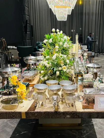 A stunning buffet island at a private event, featuring elegant silverware, chafing dishes, and a towering floral centerpiece. This is how we make our food stations a focal point of the decor.