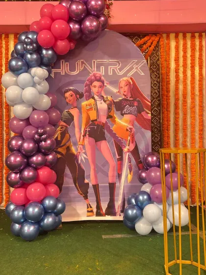 A 'Huntrix' themed party setup with a balloon arch in shades of purple, pink, and blue, set against a backdrop of marigold garlands.