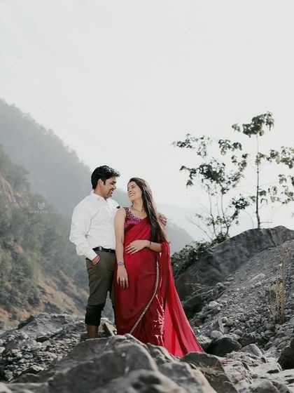A romantic portrait of a couple in a rocky, natural landscape. The woman's red saree provides a stunning pop of color against the earthy tones.