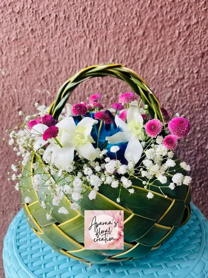 A close-up of a finished coconut leaf basket, filled with white, blue, and purple flowers. The texture of the woven leaves adds a rustic and traditional feel.