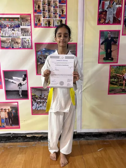 A young student proudly holds her certificate after successfully passing her taekwondo belt test. The wall behind her is filled with photos of other students, showcasing our growing community of martial artists.