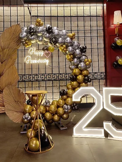 An elegant 25th-anniversary decoration for a milestone celebration. This setup features a balloon arch in black, silver, and gold, a "Happy Anniversary" neon sign, and large light-up numbers.