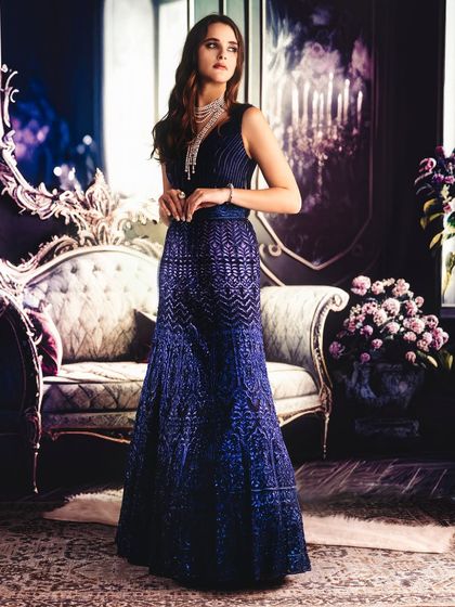 A luxurious, moody interior is projected to create a sophisticated atmosphere. The lighting is soft and directional, highlighting the texture of the blue gown and creating a sense of quiet elegance.