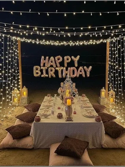 A unique and romantic beach dinner setup, with a low table, cushions, and a canopy of fairy lights creating an unforgettable birthday experience.