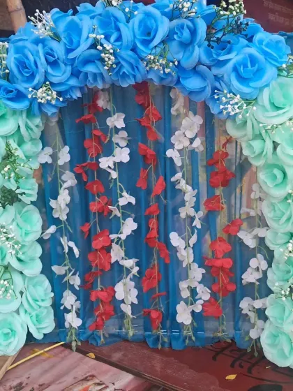 A cool-toned color palette for a festival backdrop. This design uses an arch of blue and sea-green artificial roses over a blue curtain, with contrasting red and white floral strings hanging in the center.