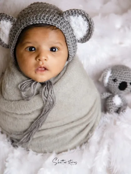 Hello, little koala! This newborn is wrapped in grey and wearing an adorable crocheted koala hat, looking right at the camera.