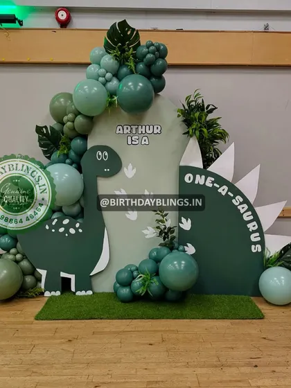 A "One-a-saurus" first birthday party with a cute, cartoon dinosaur theme. The setup uses a custom-shaped backdrop to mimic a dinosaur's back.