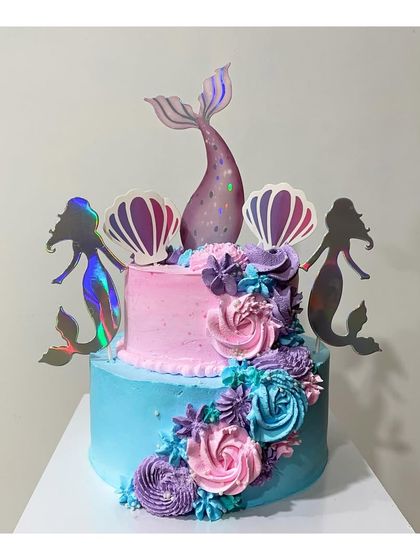 A beautiful 2-tier mermaid theme cake. The design uses shades of pink, purple, and blue, with iridescent toppers to create a magical under-the-sea look.