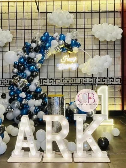 For a classy and simple first birthday, we designed this cloud-themed decor. The setup includes a balloon arch in shades of blue, black, and silver, marquee letters spelling the name 'ARK', and soft cloud-like balloon clusters to create a dreamy atmosphere.