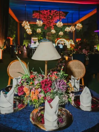 Every table setting is an opportunity for beauty. This centerpiece features a vibrant floral arrangement with a unique table lamp, set on a deep blue velvet tablecloth with ornate charger plates.