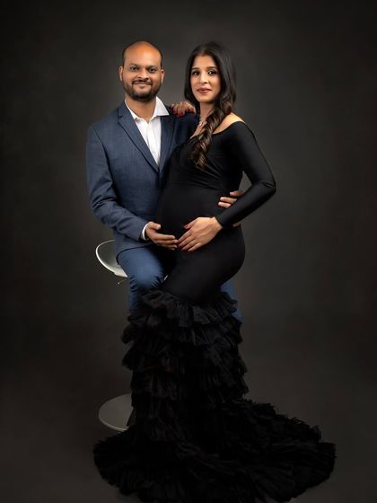 A sophisticated couple portrait in the studio. The mum-to-be is seated on a stool while her partner stands beside her, creating a balanced and elegant composition.
