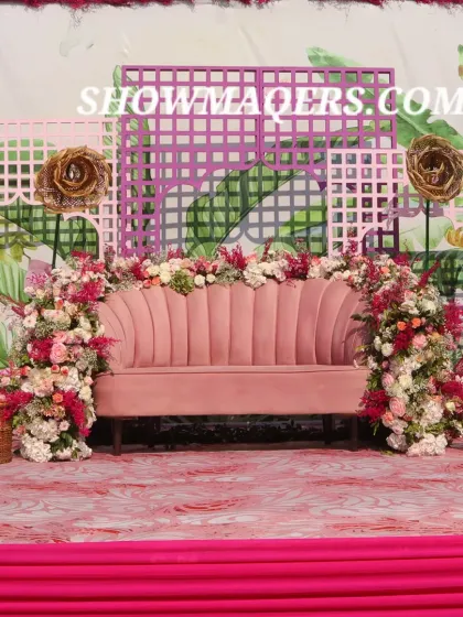 The main seating stage for the couple at the Haldi, featuring a plush pink sofa set against a tropical print backdrop and surrounded by flowers.