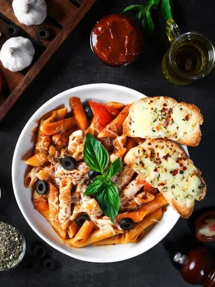 A top-down, moody shot of a bowl of Arrabiata pasta with garlic bread. The dark background and scattered ingredients like olives and herbs create a dramatic and sophisticated image.