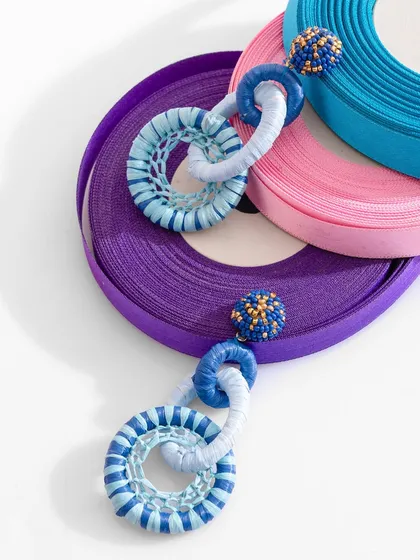 A creative flat-lay of blue and white chain-link earrings on spools of ribbon. The composition is playful and colorful, showcasing the earrings' unique design in an artistic way.