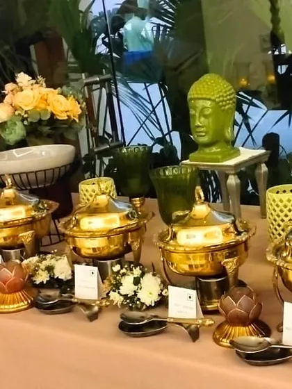 A daytime buffet setup showcasing a mix of modern ceramic bowls and traditional brassware. The decor includes vibrant floral bouquets and a serene Buddha head statue, reflecting a tranquil theme.