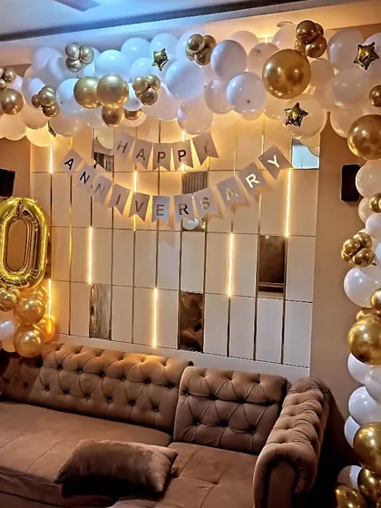 A beautiful home decoration for a 20th anniversary. The living room is transformed with a 'Happy Anniversary' banner, large number balloons, and a flowing white and gold balloon arch.