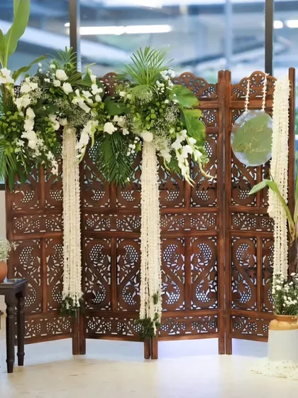 I designed this elegant and rustic backdrop for Vivek and Maria's engagement ceremony. The setup features a beautifully carved wooden screen, adorned with fresh white flowers, lush greenery, and traditional Indian decor elements.