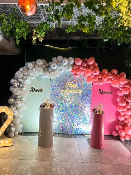 A personalized anniversary setup with a sparkling silver sequin wall. The decor includes two backdrops with the couple's names and balloon arches in coordinating silver and pink.