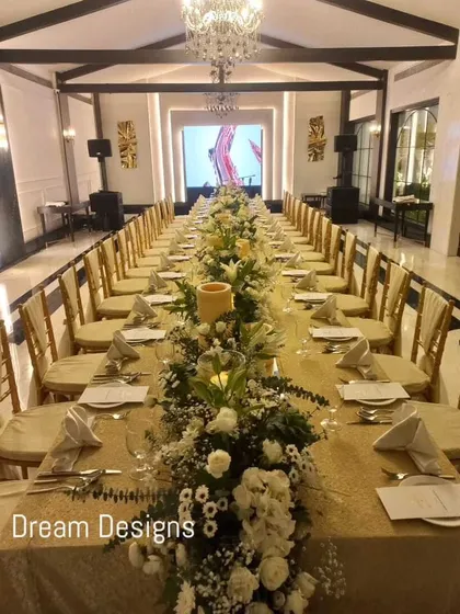A formal sit-down dinner setup inside a grand hall. A long table is beautifully decorated with a continuous runner of white flowers and greenery, creating a warm and inviting atmosphere for a celebratory meal.