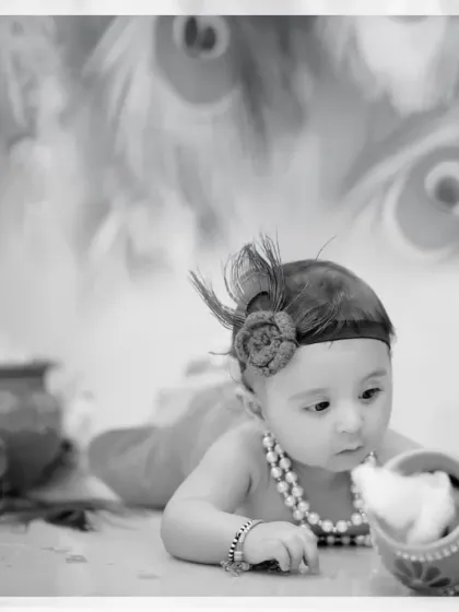 A soft and artistic black and white photo of a baby dressed as Krishna, capturing a moment of quiet curiosity.