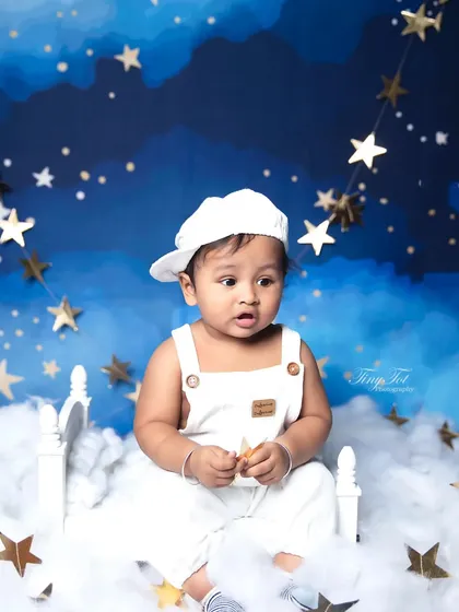 A curious expression captured during a starry night themed photoshoot. The baby is sitting on a tiny bed amidst fluffy clouds, creating a whimsical portrait.