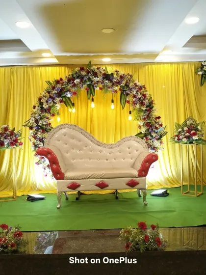 A reception stage with a bright yellow backdrop and a multi-colored floral arch. The classic red and white diwan adds a touch of traditional elegance to the vibrant setting.