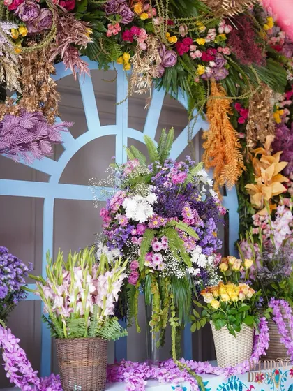An abundant floral arrangement in shades of purple, white, and yellow, set against a light blue window frame. This image showcases the sheer volume and variety of flowers used to create a lush, celebratory feel.