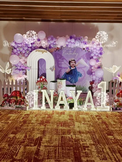 A beautiful fairy-themed first birthday stage for Inaya. The setup includes a backdrop with a photo of the birthday girl, marquee letters, a white picket fence, and whimsical mushroom props.