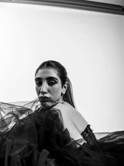 This shot includes the studio lighting equipment in the frame, giving it a raw, behind-the-scenes feel. The black and white treatment enhances the graphic quality of the composition, blending the model's elegance with the industrial feel of the studio.