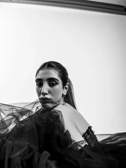 This shot includes the studio lighting equipment in the frame, giving it a raw, behind-the-scenes feel. The black and white treatment enhances the graphic quality of the composition, blending the model's elegance with the industrial feel of the studio.
