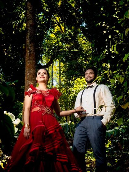 A creative pre-wedding photo taken in a lush, green forest. The couple's outfits, a red gown with illusion details and a vintage-style shirt with suspenders, create a unique look.